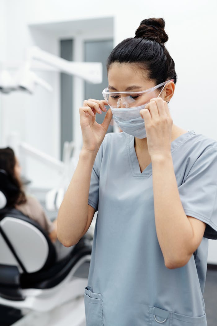 Asian dentist in scrubs adjusting face mask and goggles in a dental office. Professional healthcare scene.